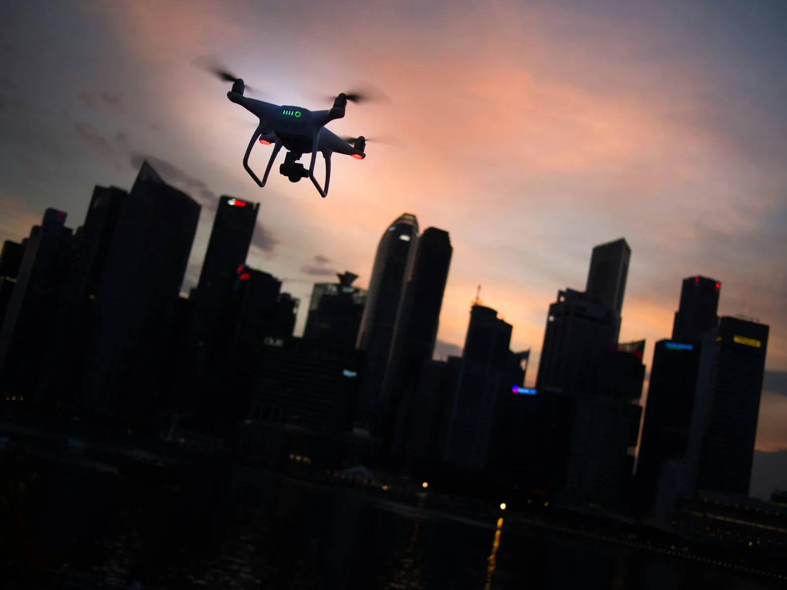 The silhouette of a quadcopter drone hovering against a lit city skyline at dusk.